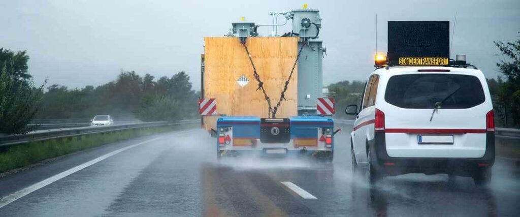 An escort vehicle driving behind a semi transporting an oversized load in rainy weather conditions